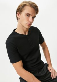 Camiseta henley de manga corta negra con tejido texturizado, tapeta de botones y escote redondo, combinada con pantalones negros sueltos.
