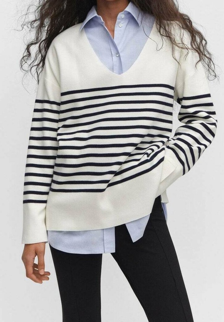 Personne portant un pull blanc à rayures horizontales bleu marine par-dessus une chemise bleu clair boutonnée et un pantalon noir, main dans la poche.