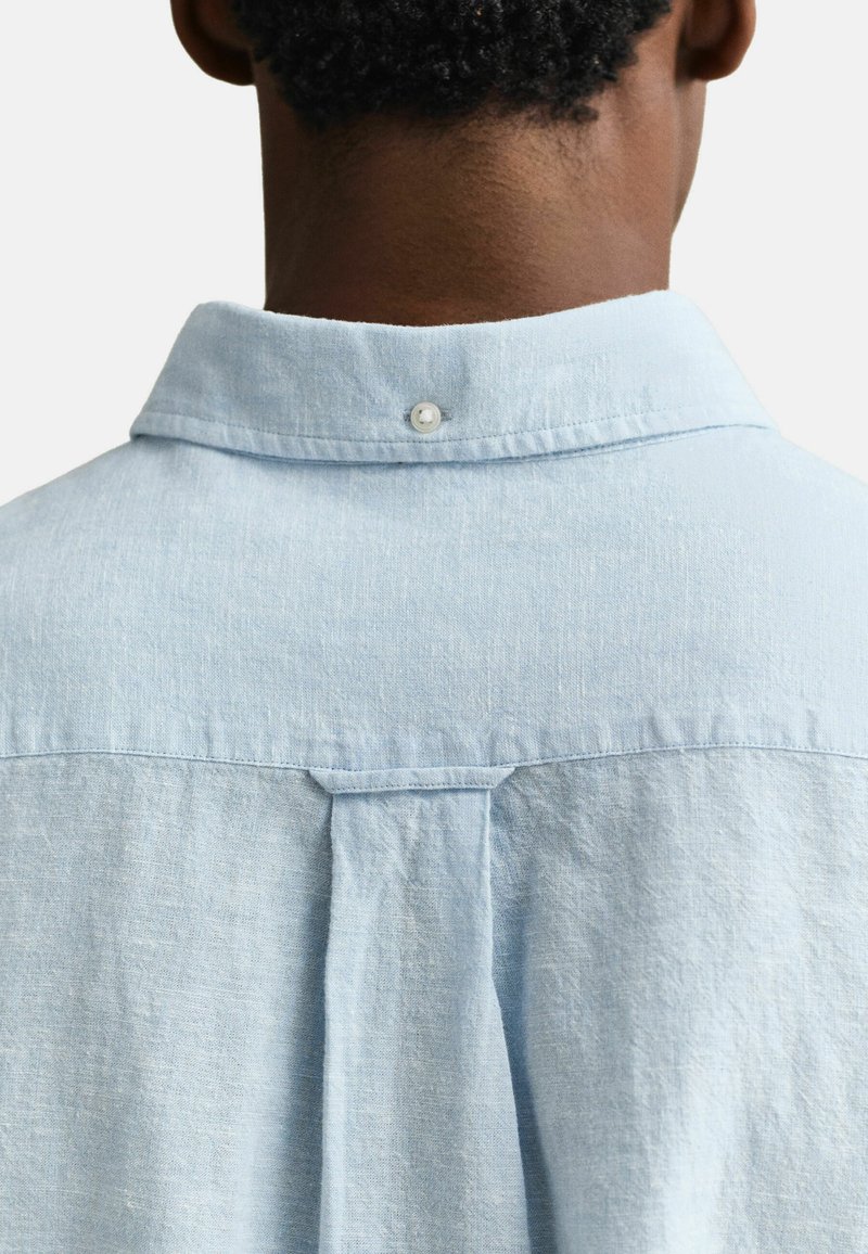 Set bagfra af en person iført en lyseblå hørskjorte med en knappet kraveflip og en central kasseplissé på den øvre ryg.