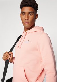 Haut à capuche rose avec une poche kangourou, capuche à cordon de serrage ajustable et logo Puma noir sur la poitrine. Texture lisse et coupe décontractée.