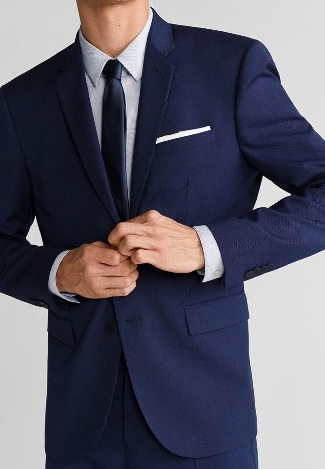 Homme boutonnant une veste de costume bleu marine sur une chemise bleu clair et une cravate foncée, avec un mouchoir blanc dans la poche, sur un fond uni.