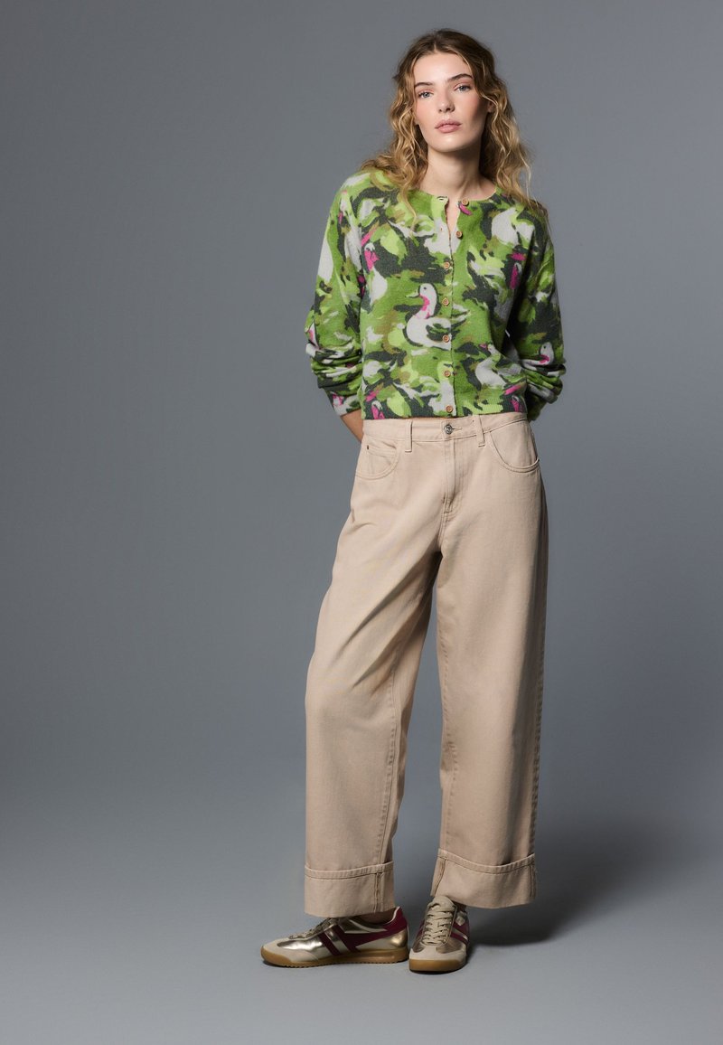 Jeune femme debout, les mains dans les poches, portant un cardigan camouflage vert, un pantalon large beige et des baskets dorées sur fond gris.