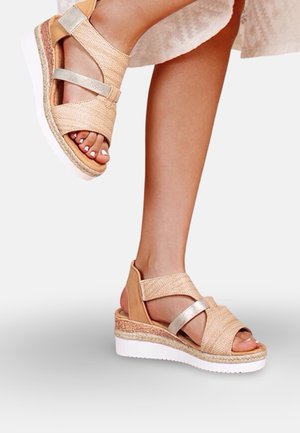 Femme portant des sandales compensées beige et métalliques avec une texture tissée et des semelles blanches, debout avec un pied légèrement levé.