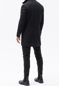 Svart lång kappa med hög krage, parat med åtsittande svarta byxor och eleganta ankelboots. Slät tyg med skräddarsydd design och minimal hårdvara.