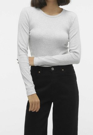 Vrouw draagt een lichtgrijs, nauwsluitend shirt met lange mouwen en een zwarte broek met hoge taille tegen een effen witte achtergrond.