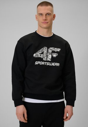 Młody mężczyzna z krótkimi włosami w czarnej bluzie z białym, graficznym logo "4F Sportswear" z przodu, na jednolitym szarym tle.