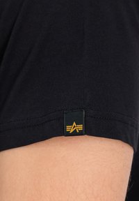 T-shirt en coton noir à manches courtes, avec une étiquette en bas en vert foncé et or comportant un logo et trois lignes horizontales.