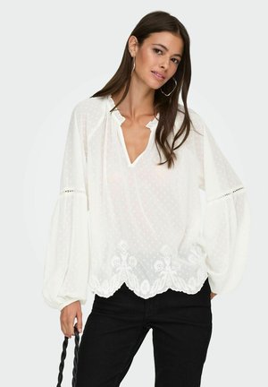 Vrouw met lang bruin haar, gekleed in een witte doorschijnende blouse met opgezette mouwen en bloemig borduurwerk, die een gedraaide zwarte tas hengsel vasthoudt.