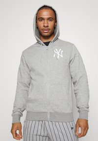 Sudadera gris con cremallera y capucha, que presenta un logo blanco en el lado izquierdo del pecho y bolsillos delanteros. La tela parece suave y tiene puños y dobladillo acanalados.