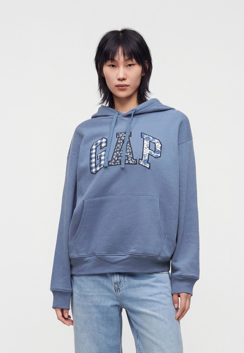 Personne portant un sweat à capuche GAP bleu avec des lettres à motifs et un jean bleu clair, debout devant un fond blanc uni.