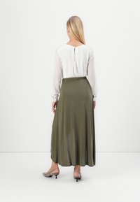 Langer olivgrüner Rock mit einer leichten, fließenden Textur, kombiniert mit einer weißen Bluse, die durchsichtige gestreifte Ärmel und einen Knopfverschluss auf der Rückseite aufweist.