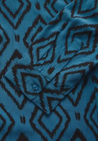 Tessuto blu a motivo con un design a zigzag nero, con un primo piano di un polsino abbottonato e una superficie liscia.