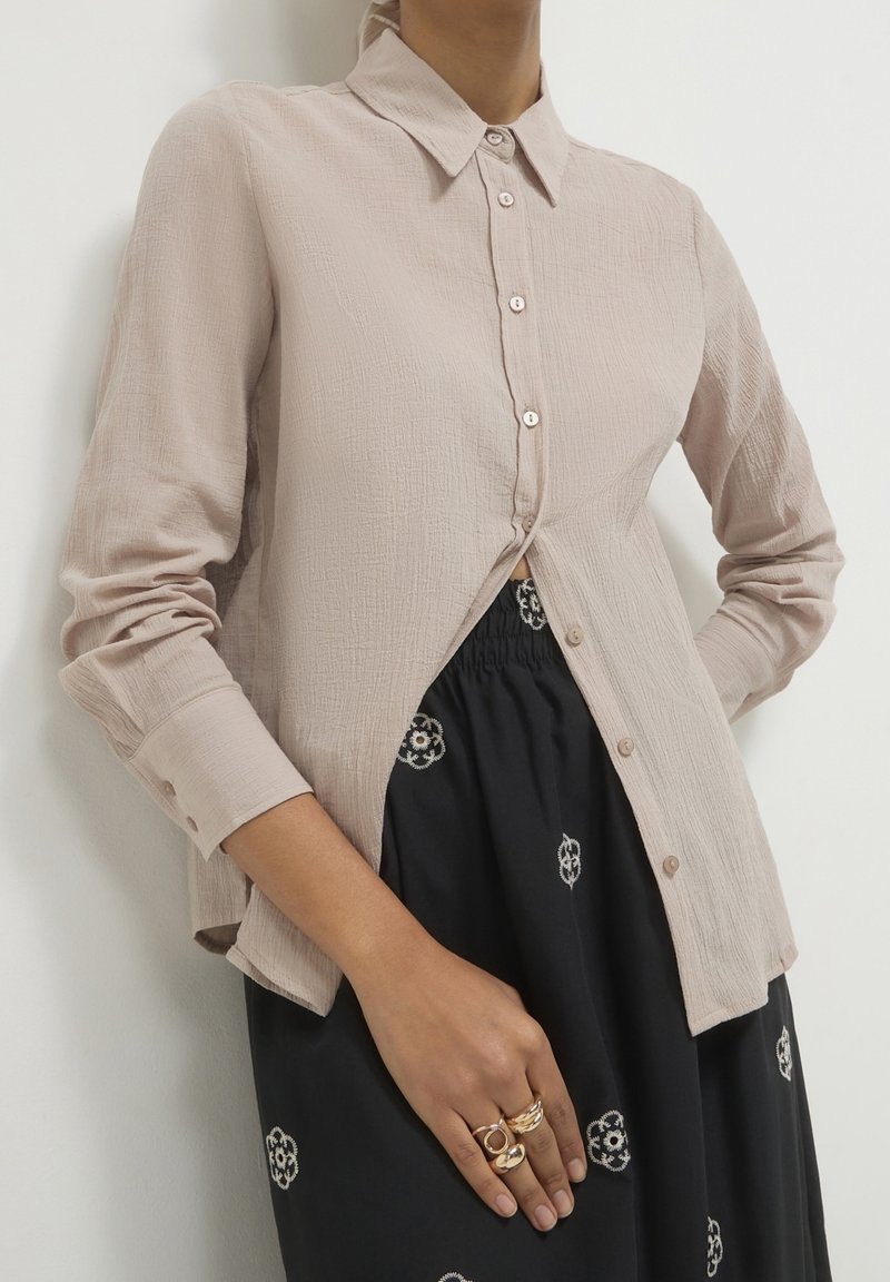 Femme portant une chemise beige texturée avec boutons et une jupe noire à motifs floraux, montrant plusieurs bagues en or sur sa main reposant sur la jupe.