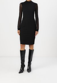 Schwarzes Kleid mit tailliertem Oberteil, hohem Kragen und durchsichtigen langen Ärmeln. Getragen mit eleganten schwarzen kniehohen Stilettos. Glatter Stoff.