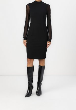 Robe noire avec un corsage ajusté, un col haut et des manches longues en voile. Associée à des bottes noires à talons aiguilles et montantes jusqu'au genou. Tissu lisse.