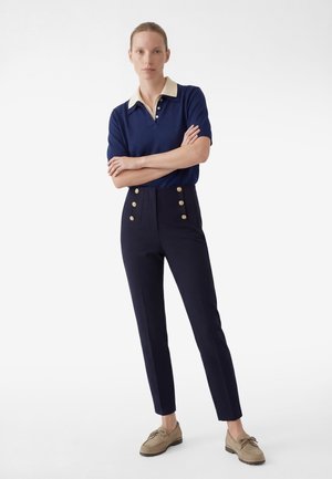 Mujer de pie con los brazos cruzados, vestida con una camisa polo de manga corta azul marino y pantalones de talle alto azul marino con botones dorados, y mocasines beige.