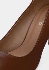 Καφέ δερμάτινο παπούτσι με ψηλό τακούνι και στρογγυλεμένη μύτη, με λεία υφή και γυαλιστερό φινίρισμα. Το εσωτερικό έχει ανοιχτότερο καφέ χρώμα.
