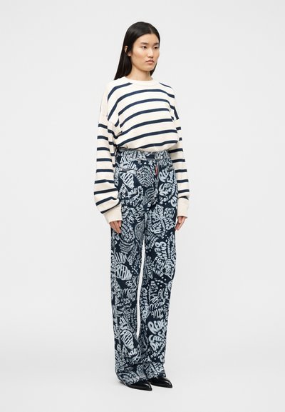 Ριγέ πουλόβερ σε κρεμ χρώμα με άνετη εφαρμογή. Παντελόνι με φαρδιά μπατζάκια σε ναυτικό μπλε με λευκή γραφική επιγραφή. Το μοντέλο φοράει μυτερά μαύρα παπούτσια.