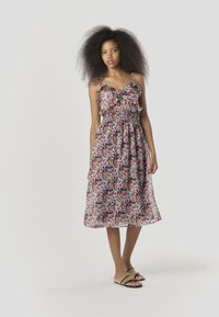 Kleid mit Blumenmuster, gerafften Trägern, geraffter Taille und halbtransparentem Stoff. Die Farben sind Pink, Blau und Grün. Kombiniert mit schwarzen Sandalen.