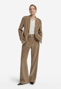Braune Cord-Jacke und weite Hosen mit strukturiertem Finish. Kombiniert mit einem weißen Polo und einem geflochtenen Gürtel, dazu spitz zulaufende schwarze Schuhe.