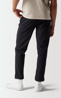 Pantalones negros hechos de una tela suave y ligera con un diseño ajustado. Llevados con zapatillas blancas de caña alta. Fondo neutro.