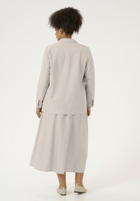 Lichtgrijze pinstripe blazer met een achtervent, gecombineerd met een bijpassende lange rok. De look wordt afgemaakt met beige platte schoenen.