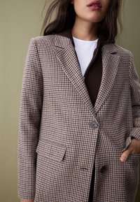 Brauner und beiger Blazer im Hahnentritt-Muster mit Reverskragen, einem Knopfverschluss und seitlichen Taschen. Über einem dunkelbraunen Oberteil getragen.