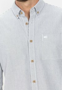 Chemise à rayures en coton gris et blanc avec boutons. Elle dispose d'un col classique, de boutons en bois et d'une poche plaquée avec un logo.