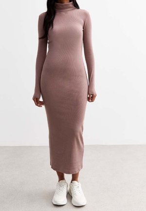 Femme portant une robe à col roulé côtelée mauve à manches longues, associée à des baskets blanches, debout devant un fond clair uni.