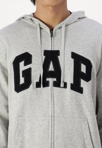 Hellgrauer Zip-Hoodie aus Stoff mit weicher Textur. Verfügt über eine große schwarze "GAP"-Applikation auf der Brust und Kordeln an der Kapuze.