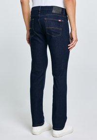 Bărbat purtând blugi drepti albastru închis cu buzunare pe spate, adidași albi și un tricou alb, stând în fața unui fundal simplu.