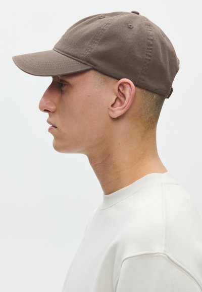 Ung mand i hvid skjorte iført brun baseballkasket, vist i profil vendt mod venstre mod en ensartet lys baggrund.