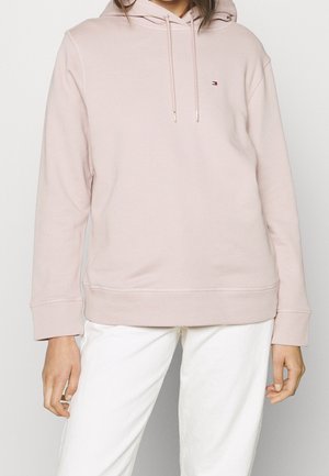 Henkilö, joka pukeutunut vaaleanpunaiseen huppariin, jossa on pieni logo rinnassa, ja valkoisiin housuihin, seisoo yksiväristä taustaa vasten.