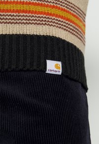 Pull beige tricoté avec des rayures orange, jaune et marron au-dessus d'un ourlet côtelé noir et une étiquette logo Carhartt blanche.