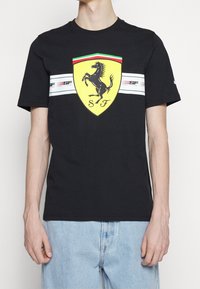 Svart bomull t-shirt med en gul Ferrari-logotyp och en prancing horse-design. Har vita horisontella ränder med röda och gröna detaljer.