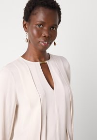 Lys beige bluse med nøkkelhull-hals, vertikale folder og en gullakcent nær kragen. Stoffet ser mykt og glatt ut.