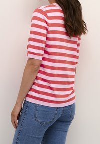 Mujer con cabello castaño largo, vistiendo una blusa de mangas cortas a rayas rosa y roja, y jeans azules, de pie frente a una pared clara y sencilla.