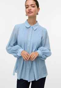 Lichtblauwe blouse van textuurstof, met een kraag, knopen aan de voorkant en opgezette lange mouwen met gerimpelde boorden.