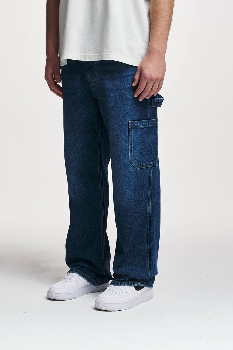 Prostorne temno modre denim hlače z velikim stranskim žepom, z detajli šivanja in sproščenim krojem, v kombinaciji z belimi teniskami.