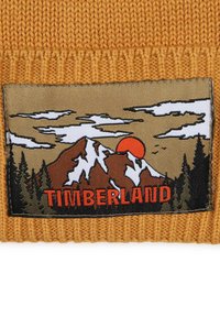 Bonnet en tricot orange présentant une étiquette en tissu rectangulaire avec un paysage de montagne, un soleil, des nuages et "TIMBERLAND" en lettres orange en gras.