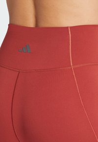 Leggings rouges taille haute avec une texture lisse, arborant un logo sombre près de la taille et des coutures piquées pour un détail supplémentaire.