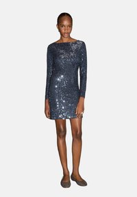 Morska haljina sa šljokicama, dugih rukava, prilagođenog silueta i izrezom u obliku uvale; model nosi ravne, točkaste cipele za kontrast.