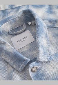 Light blue denim shirt with faded patterns, a classic collar, and a silver button featuring a logo, labeled with "LES DEUX."