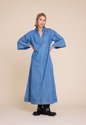 Kvinne stående med hendene på hoftene, iført en blå lang denimkjole og svarte ankelstøvler foran en beige bakgrunn.