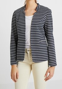 Blazer i marinblått med vita horisontella ränder, med en klädd krage, två framfickor och en lätt tygstruktur.