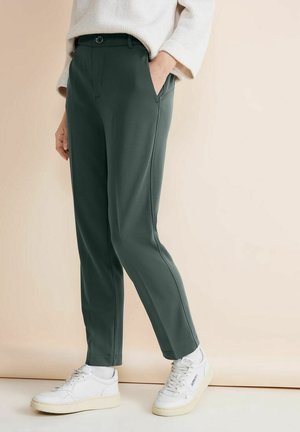 Pantalon vert sur mesure avec une texture lisse, équipé de poches latérales et d'une ceinture classique. Associé à des baskets blanches.