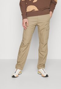 Beige cargobyxor med flera fickor, tillverkade av slitstarkt material, har en avslappnad passform och raka ben, i kombination med vita sneakers.