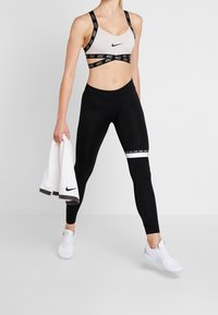 Kvinna i svart och vit Nike sport-bh, svarta leggings med vit rand, vita sneakers, hållande en vit handduk med Nike-logotyp.