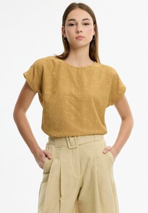 Joven con cabello castaño liso, lleva pendientes de aro, una camiseta de manga corta texturizada de color mostaza y pantalones beige plisados de cintura alta.
