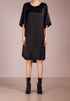 Femme portant une robe ample en satin noir longueur genou avec de larges manches et des bottines noires assorties sur un fond uni.